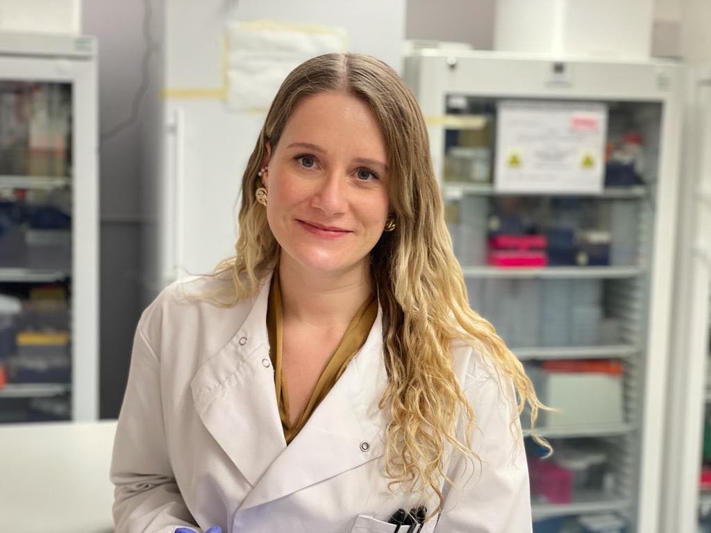 Dr Ashley Boyle, a woman with blonde wavy hair and wearing a white lab coat, smiles at the camera