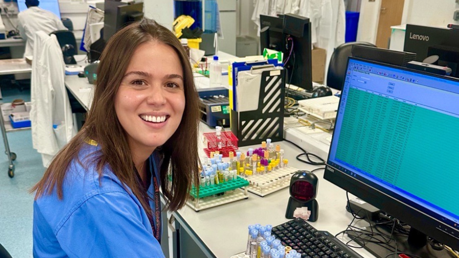 Dr Eva Ioannidou smiling at her workstation in a clinical laboratory, wearing blue scrubs, 