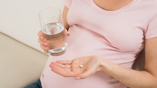 Pregnant woman holding a glass of water with a supplement tablet in her other hand.