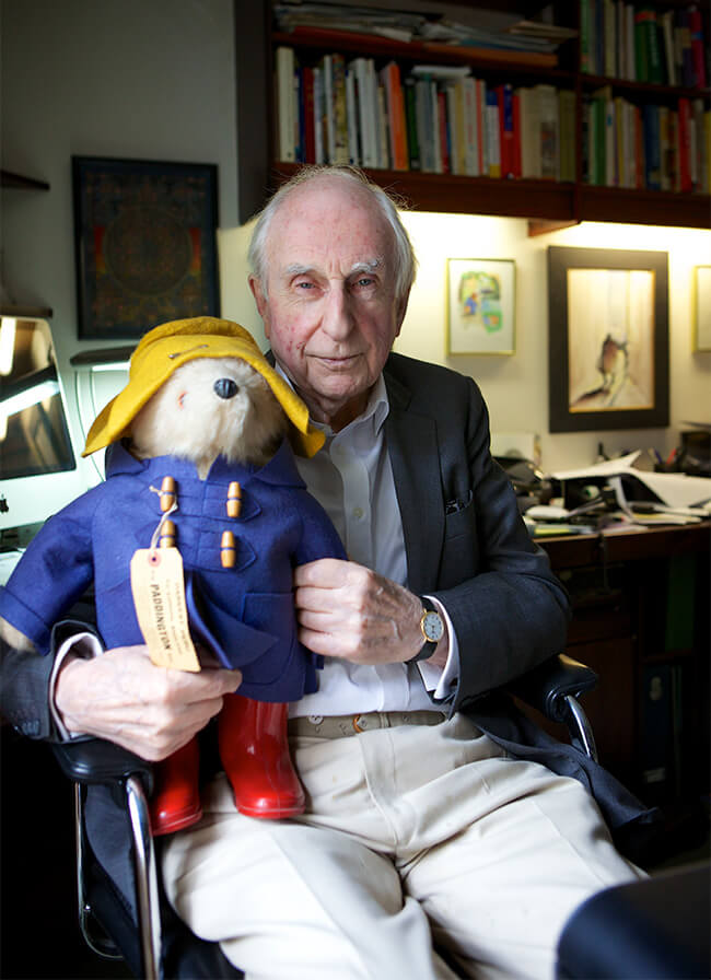 Michael Bond sitting in his study holding a Paddington Bear toy dressed in a yellow hat, blue coat, and red boots.