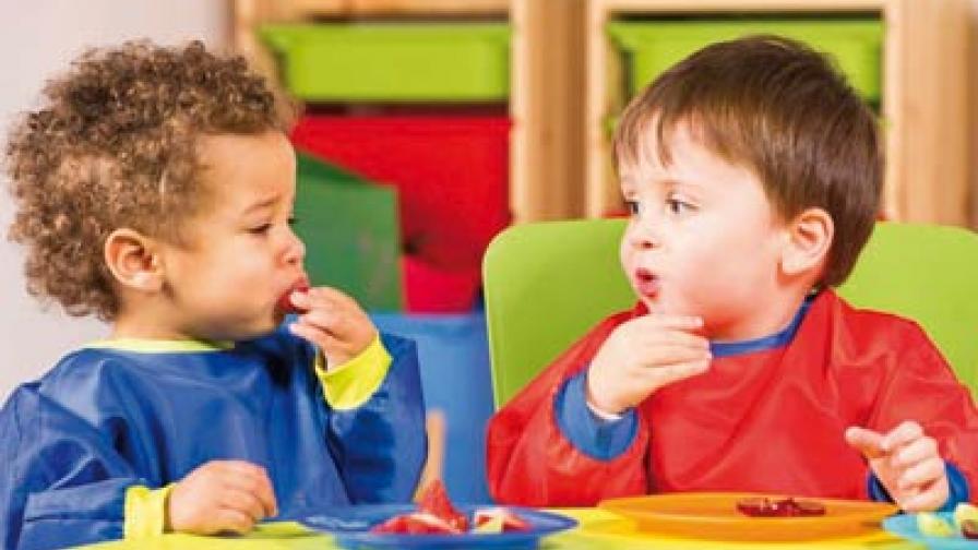 Two toddlers sitting at a table eating from a plate of fruit
