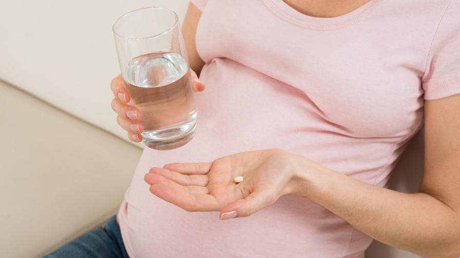 Pregnant woman holding a glass of water in one hand and a supplement tablet in her other outstretched palm.