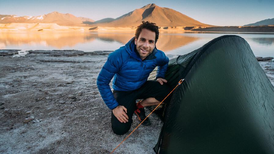 Mark kneeling beside a tend with a lake and mountain in the background