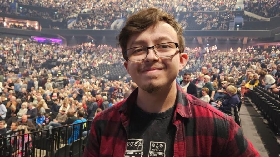 Joey now, aged 18, smiling, at a music concert venue with lots of people and seating in the background.