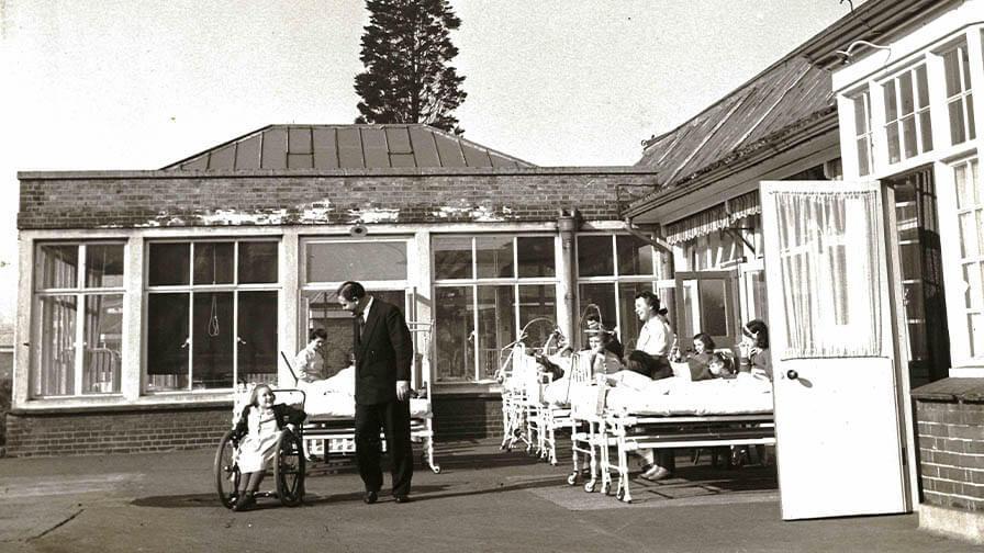 A black-and-white photo of children in wheelchairs and hospital beds outside a medical facility, with a man tending to one of them.