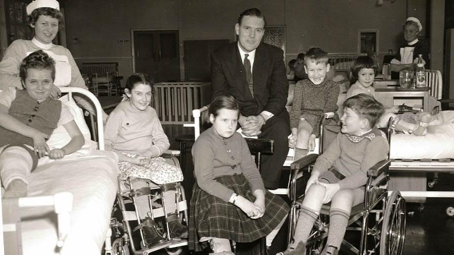 A black-and-white photo of smiling children in wheelchairs and beds at a hospital, accompanied by nurses and a man in a suit.