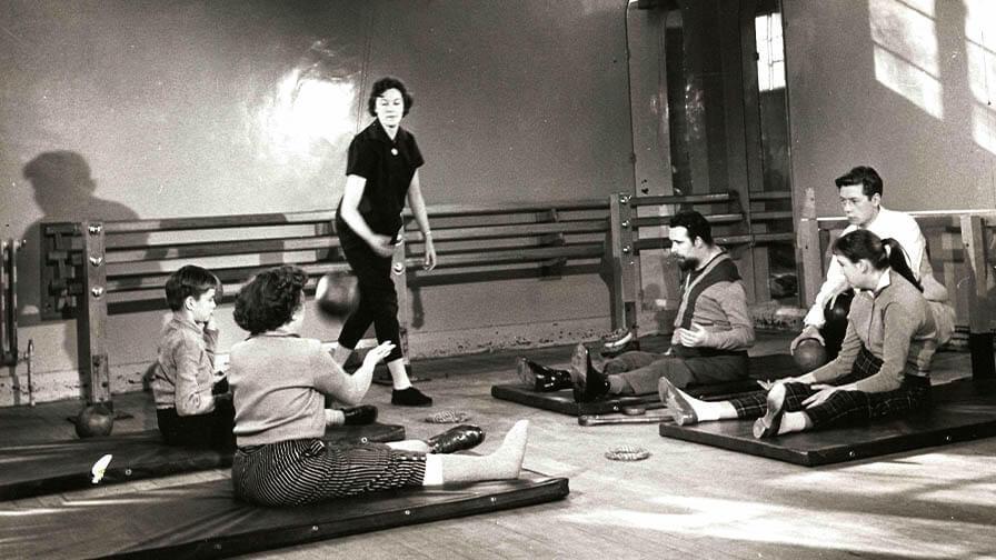 A black-and-white photo of people, including children and adults, engaging in seated physical therapy exercises, possibly polio patients.