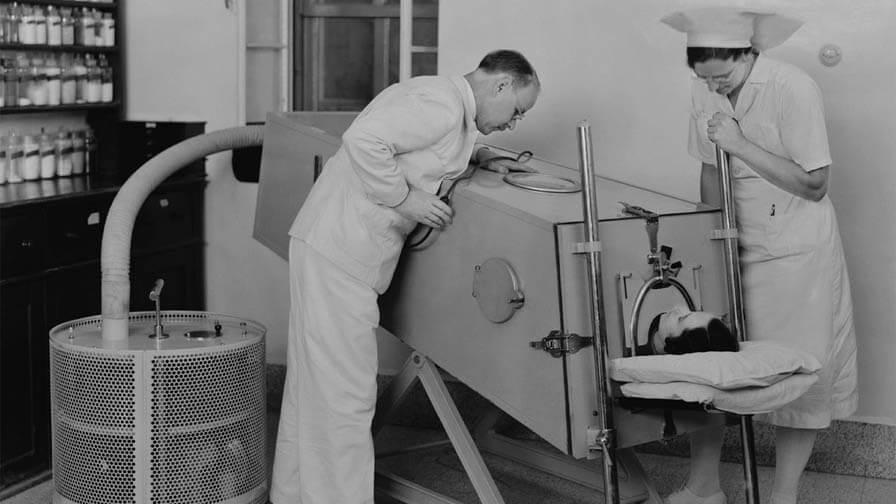A black-and-white photo of a patient in an iron lung machine, with a doctor and nurse monitoring the device in a medical setting.