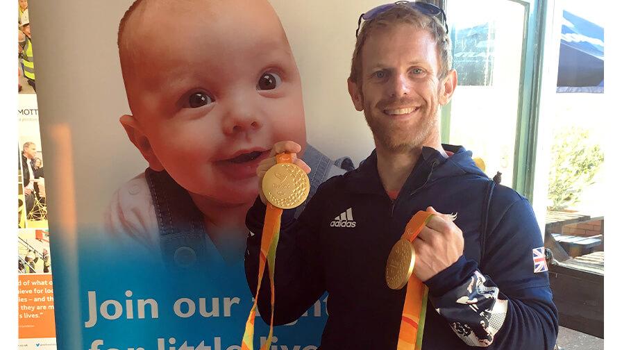 Jody Cundy CBE proudly holds two Paralympic gold medals while wearing a Team GB jacket, smiling in front of a banner supporting children.