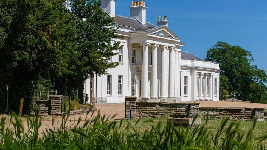 Hylands Estate, A grand white mansion with tall columns, chimneys, and a classical design, surrounded by greenery under a clear blue sky.
