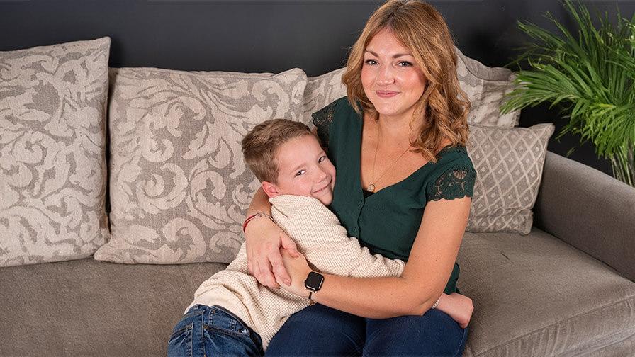 Mum Kayleigh, smiling while she sits on a sofa, embracing a young Henry with a warm, affectionate hug. Cushions and a green plant are in the background.