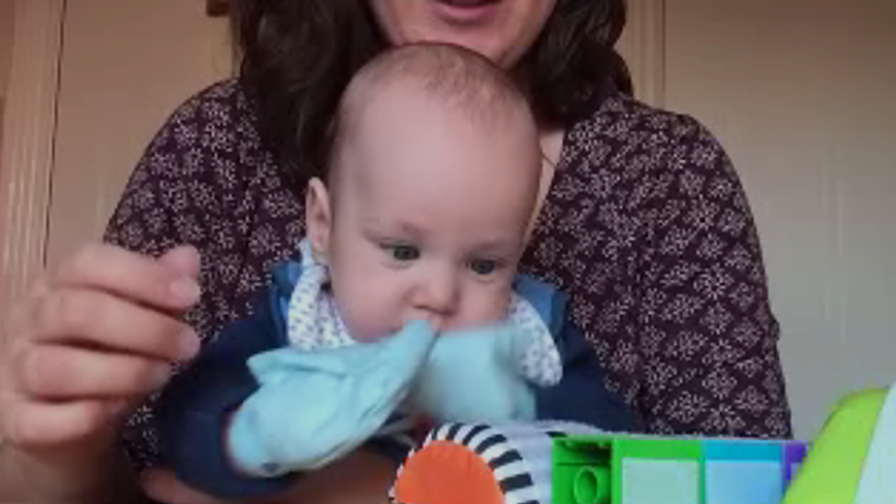 Baby wearing blue sticky mittens, playing with toys.