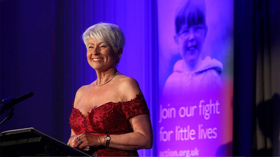 Pamela Ballantine in a sparling red dress stands behind a microphone speaking at an Action event