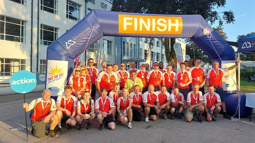 A large group of cyclists in red and white jerseys pose under a blue "FINISH" arch after completing the Race the Sun challenge.