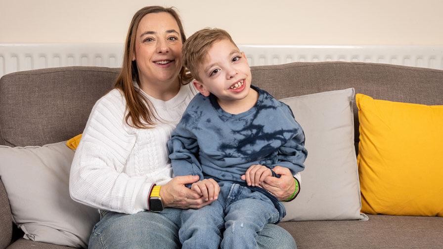 A woman and a young boy sit smiling on a gray couch with yellow and gray cushions. The woman wears a white sweater and jeans, while the boy, who appears to have a disability, wears a blue tie-dye sweatshirt and jeans. They are holding hands and looking happy.