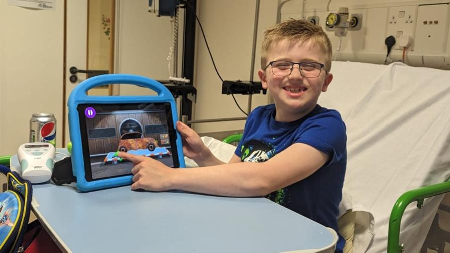 Digby, a young boy who has Duchenne muscular dystrophy. He is sitting up in a hospital bed, smiling and showing the screen of his tablet or iPad.