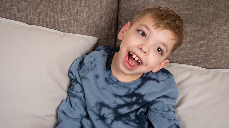 Smiling young boy in a blue tie-dye shirt sits on a couch with beige cushions, looking cheerful and engaged.