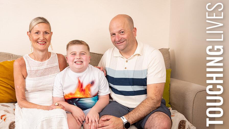 Smiling boy sits on a sofa between a woman in a white dress and a man in a striped polo, posing happily together indoors.