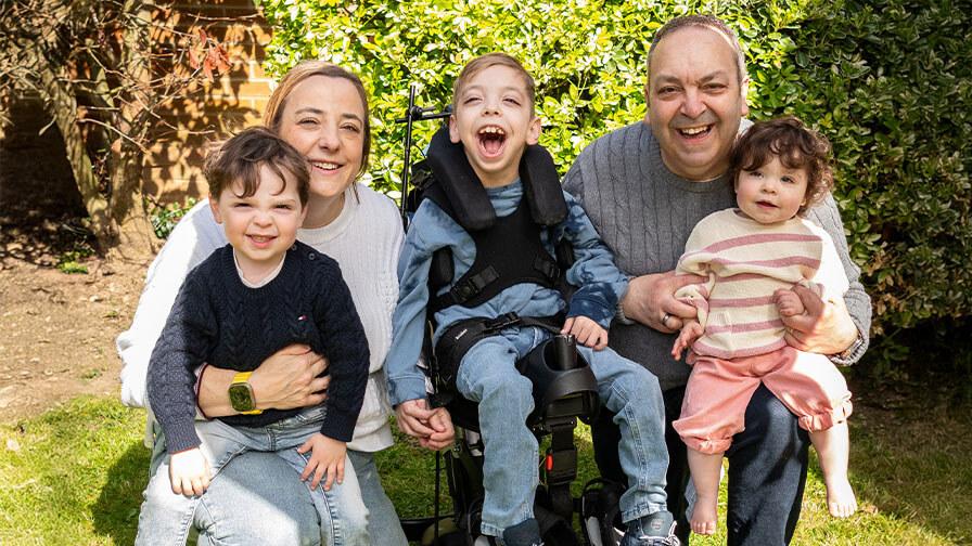 Smiling family of five, including a boy in a wheelchair, pose happily together outdoors in a sunny garden with green bushes behind them.