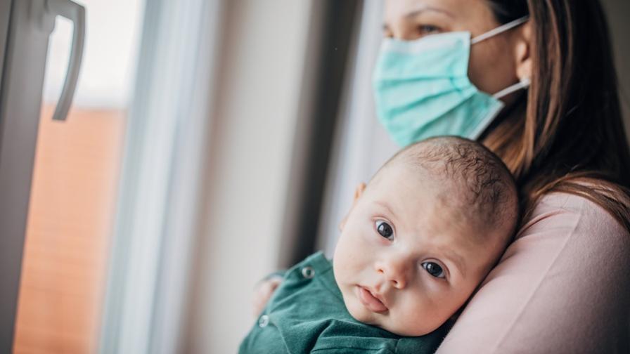 A baby, held by its mother, gazes out at the viewer. The mother is standing by a closed window and wearing a face mask over her nose and mouth.