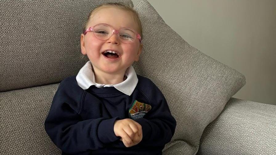 A young girl, aged about five, with Rett syndrome. She is propped up to sit on a sofa, wears glasses and is smiling.