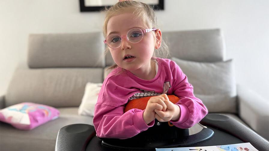 Mollie, a young girl who has Rett syndrome. She is sat in a special supportive seat with a tray in front of her. Her hands are clasped together.