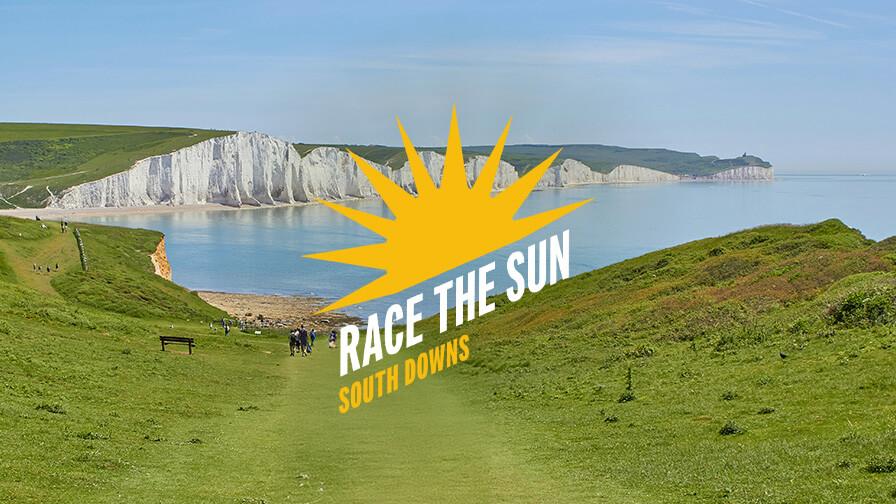 A scenic view of the South Downs coastline with white cliffs and blue sea, overlaid with a yellow sunburst logo reading "Race the Sun."