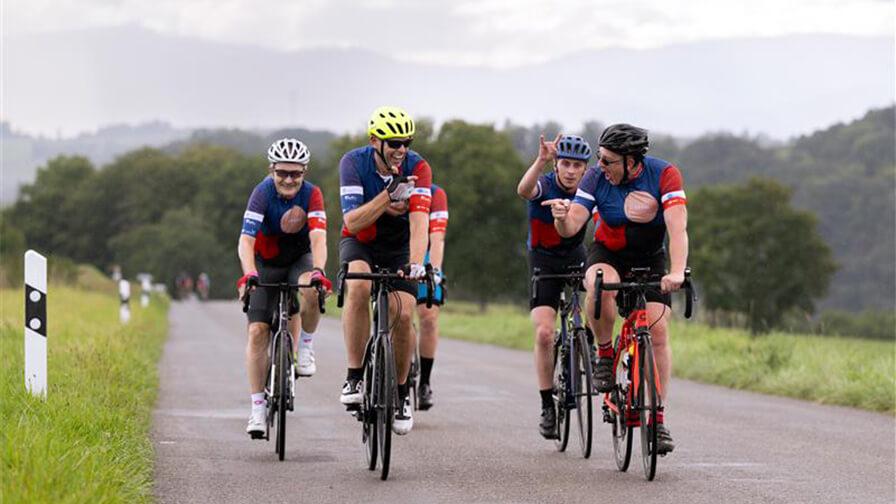 Group of cyclists wearing matching jerseys ride along a country road, chatting and smiling on a cloudy day with green hills behind them.
