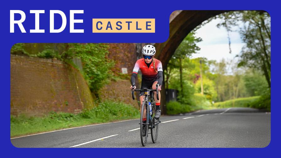 A female cyclist in a red and black jersey rides down a paved road under a brick archway.