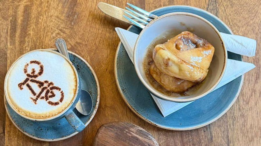 Bird’s-eye view of a coffee with bicycle latte art next to a warm cinnamon roll in a blue ceramic bowl.