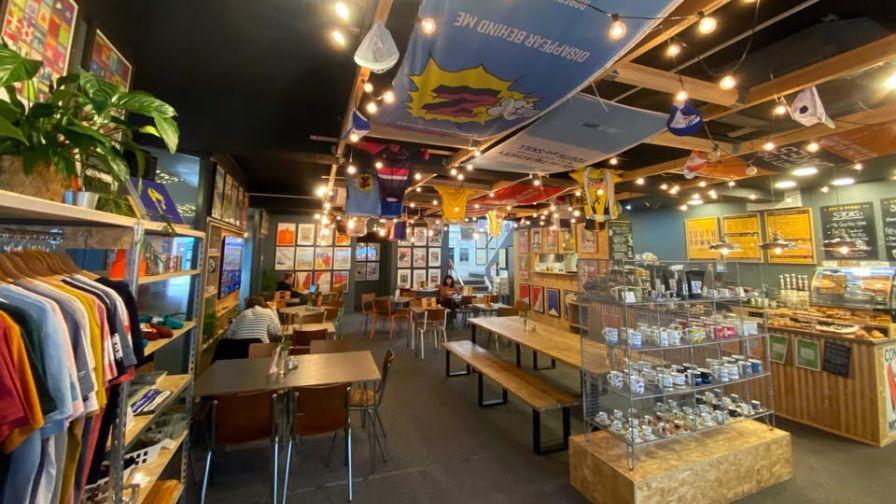 A vibrant cycling cafe filled with hanging jerseys, posters, apparel displays, and patrons at wooden tables.