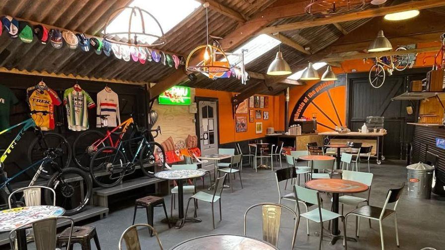 Industrial-style cafe in a workshop with orange walls, hanging bicycles, cycling caps, and metal bistro seating