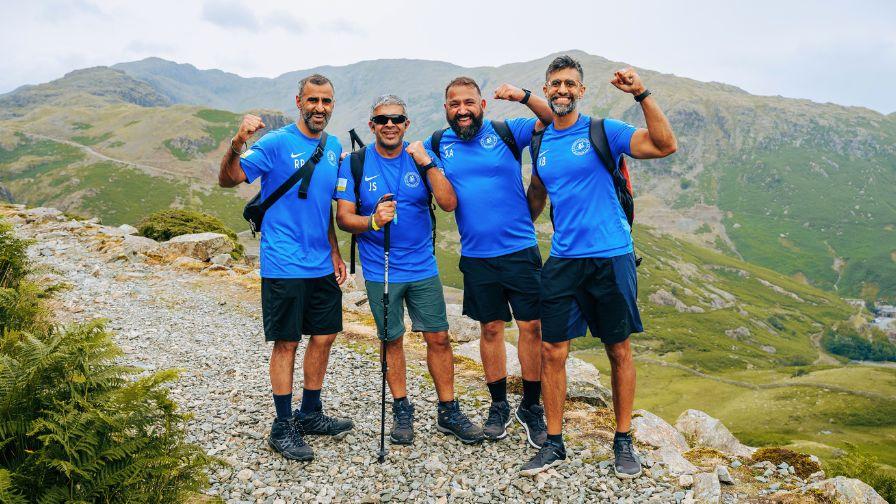 Four men in blue sports tops and hiking gear pose together on a rocky mountain path, cheering with their arms raised. The background shows a vast valley and rolling green hills under an overcast sky
