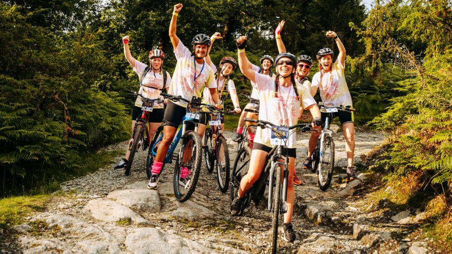 A group of women in matching team t-shirts cheering and raising their arms while mountain biking on a rocky woodland trail during the Race the Sun challenge.