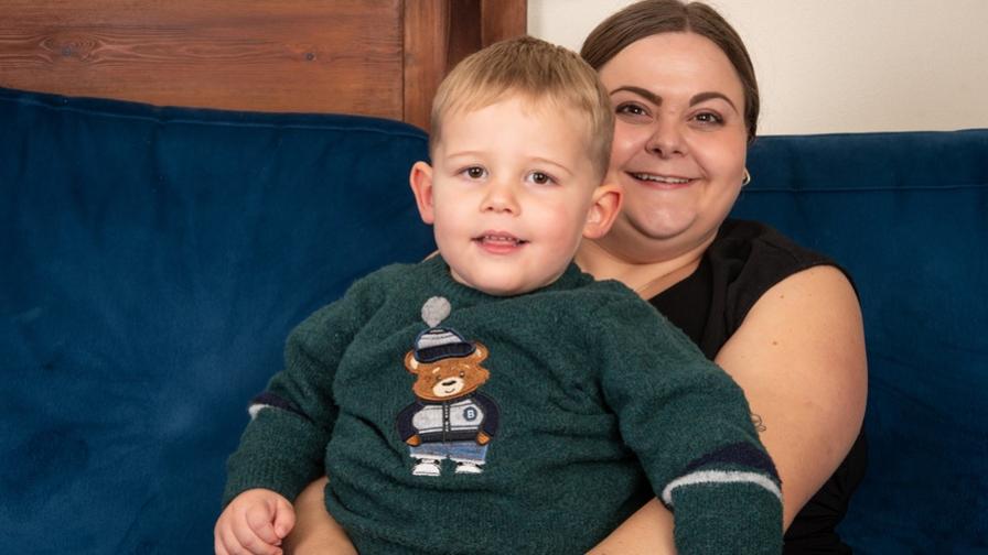 Barnaby, a young boy, with his mum. He is sitting on her lap on a blue sofa and wearing a green bear jumper.
