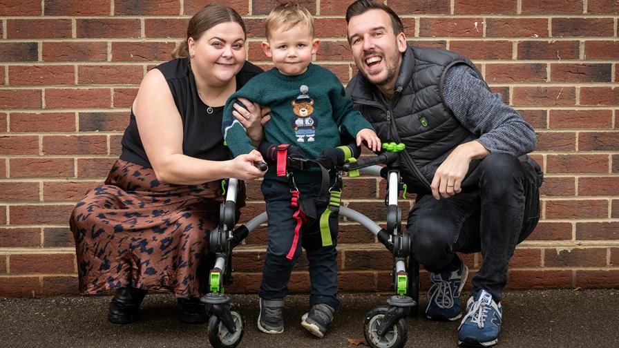Barnaby, a young boy who has severe epilepsy, with his walking frame on wheels. His mum and dad are either side of him, against a brick wall background.