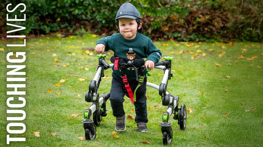 A picutre of Barnaby, a young boy in a green bear sweater and cap smiles while using a supportive walking frame in a grassy garden with fallen leaves