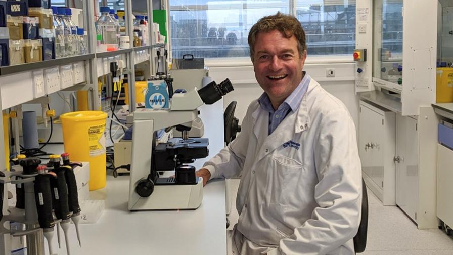 Dr Frederik van Delft sitting in a lab next to a microscope, with various medical equipment in the background. He is smiling and wearing a white lab coat.