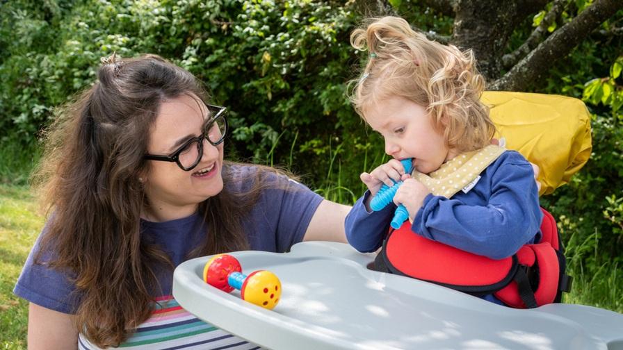 Image shows Emmy, a little girl who has Vici syndrome, with her mum, Ellie. Emmy has light brown wavy hair and wears a blue top. She is using supportive seating with a play tray in front of her. She is outside with green bushes in the background.