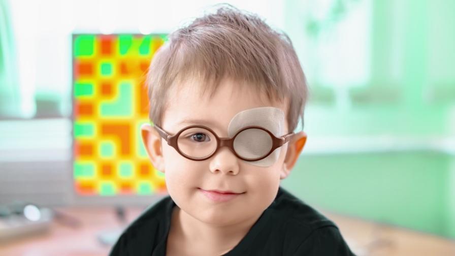 Image shows a boy, aged about two or three, wearing brown-rimmed glasses and with an eye patch over his right eye. The background is a pale green wall with a blurred window.