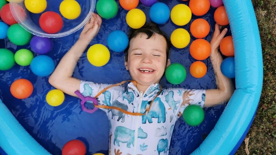 Image shows Mylo, a young boy who had CMV as a baby. He is laying, smiling, in a paddling pool filled with brightly coloured plastic balls.
