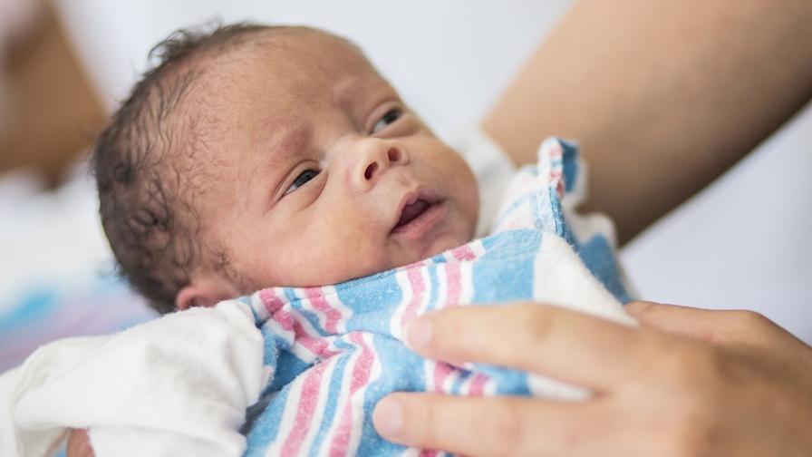 Image shows a newborn baby, with black hair, in the hands of a caregiver in a hospital setting. The baby is wrapped in a blue, pink and white stripe sheet or blanket.