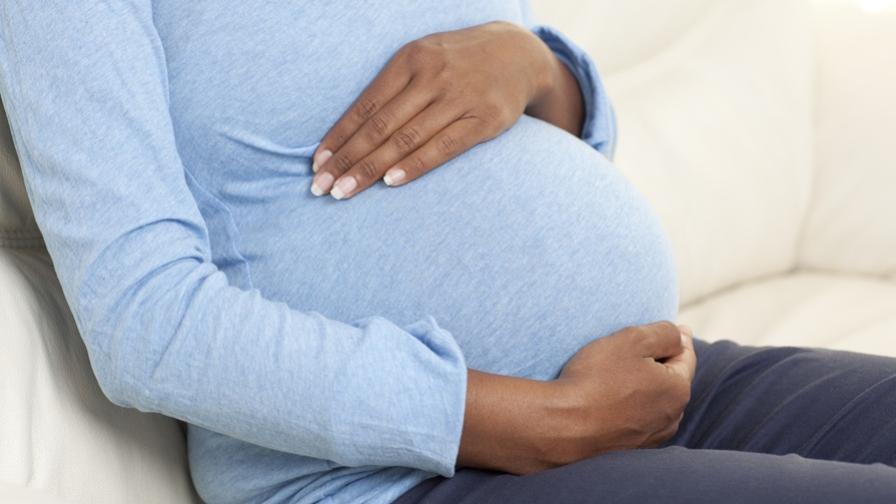 Picture of the side view of a pregnant woman, from the shoulders down, sitting on a cream sofa. Her hands are cradling her baby bump and she is wearing a blue top.