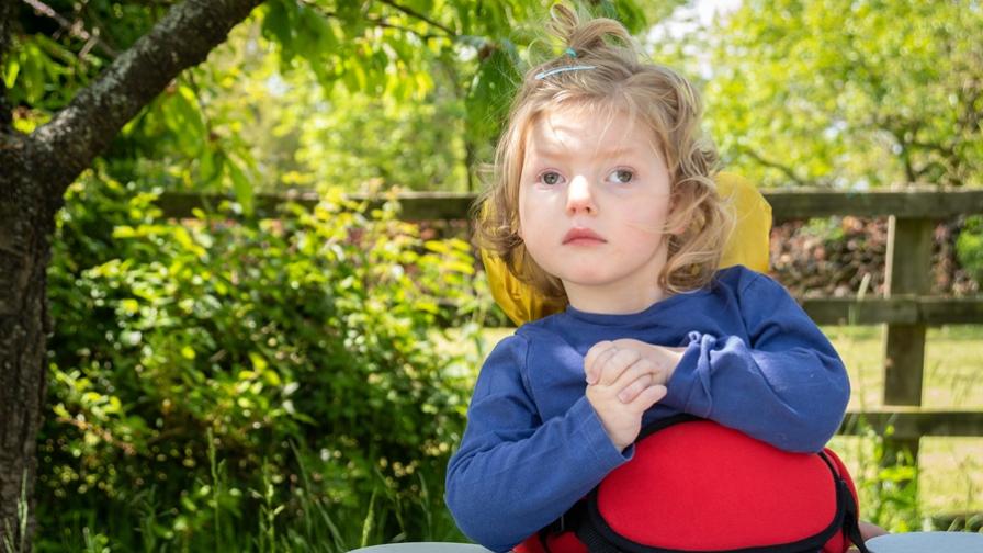 Image shows Emmy, a little girl who has Vici syndrome. She has light brown wavy hair and wears a blue top. She is using supportive seating with a play tray in front of her. She is outside with trees, bushes and a wooden fence in the background.