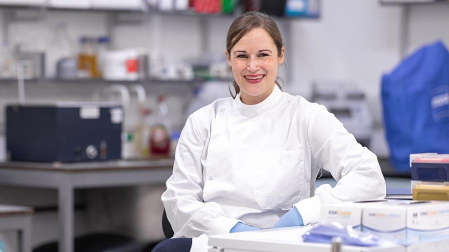 Dr Viki Male of Imperial College London, sitting in a medical research lab. She is wearing a white uniform and pale blue clinical gloves. In the background are shelves of medicine and medical equipment.