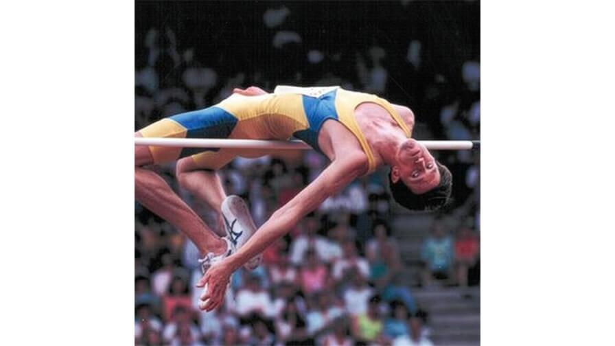 High jumper Geoff Parsons mid-air, clearing a high bar in a yellow and blue singlet during a competition with a crowd in the background.