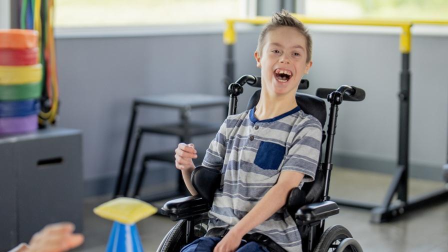 Image shows a smiling boy, aged around 12 or 13. He wearing a striped t-shirt and is in his wheelchair, with exercise equipment in the back and foreground