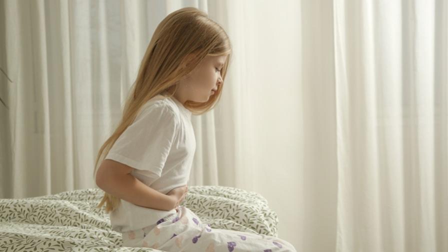 A young girl holding her stomach in pain. She has long, blonde hair and is sitting on the end of a bed, wearing pyjamas. There are cream-coloured curtains in the background.