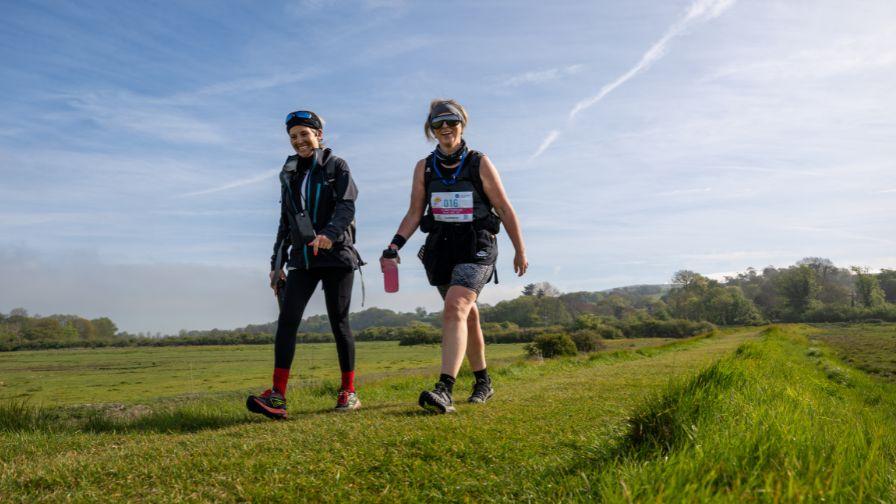 Two women wearing activewear walking along a scenic grassy trail in the countryside.