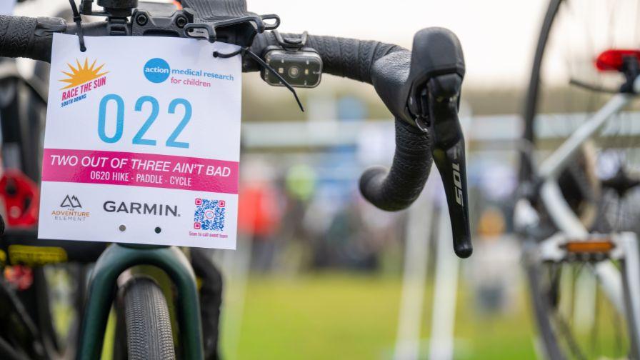 Close-up of a bicycle handlebar with an Action Medical Research event race plate numbered "022," showing a blurred outdoor event background.